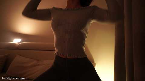 Video of a woman with fair skin, medium build, wearing a ribbed, light beige top and black pants, standing in a dimly lit bedroom with a bed and curtains.