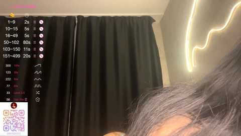 Video of a person with long, dark hair, lying on a bed with a black curtain in the background. A lightning bolt-shaped light fixture is on the wall.