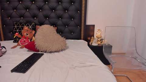 Video of a chic, modern bedroom featuring a tufted black headboard with gold accents, plush beige pillows, a red teddy bear, a clear chair, and a minimalist white desk.