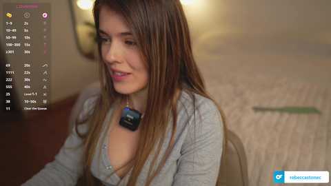 Video of a young woman with long brown hair, wearing a gray cardigan over a low-cut shirt, sitting indoors. The background features a blurred bed with a white blanket and a dimly lit room.