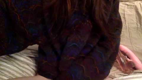 Video of a person with long, dark hair lying on a bed, their hand on their own chest. The background shows beige sheets and a pink object, possibly a phone, on the right.