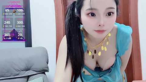 Video of an Asian woman with long black hair in a ponytail, wearing a revealing turquoise top, gold necklace, and a playful expression. Background includes a grey couch and a colorful poster.