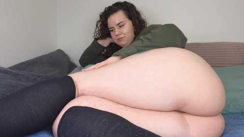 Video of a plus-size woman with curly brown hair, wearing a green sweater, black socks, and no pants, lying on a bed with grey and blue pillows, looking down.