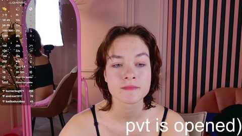 A video of a young woman with short, wavy brown hair and a neutral expression, wearing a black top, in a room with pink and black vertical striped walls.