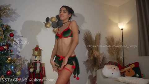 Video of a slender woman with medium skin tone in festive green and red lingerie, posing in a cozy living room with a decorated Christmas tree and holiday decorations.