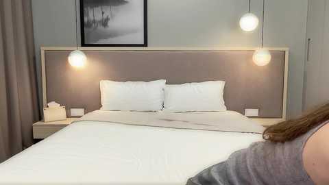 Video of a modern, minimalist bedroom with a beige upholstered headboard, two white pillows, two pendant lights, and a framed black-and-white landscape painting on the wall.