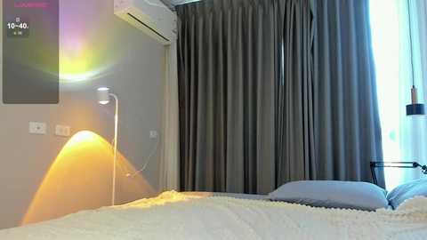 Video of a modern bedroom with a bed covered in white sheets and a grey curtain backdrop. A lamp and air conditioner are visible, casting a warm glow.