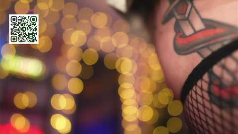 Video of a person's tattooed arm in black fishnet, with blurred bokeh lights in the background. The tattoo features a red and black design.