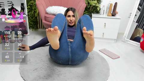 Video of a woman with long brown hair, wearing tight blue jeans, lying on her back with legs raised, in a modern, pink-tiled room with a large mirror and a pink armchair.