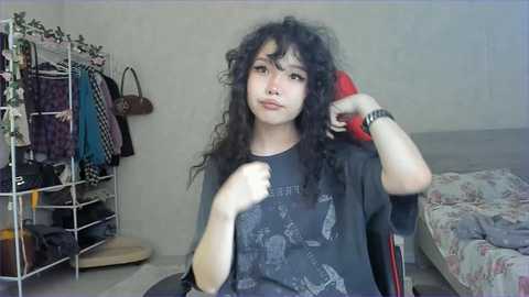 Video of a young woman with long, curly black hair, wearing a dark graphic t-shirt, sitting on a floral-patterned couch, adjusting her red headphones. Background includes a cluttered closet and beige walls.