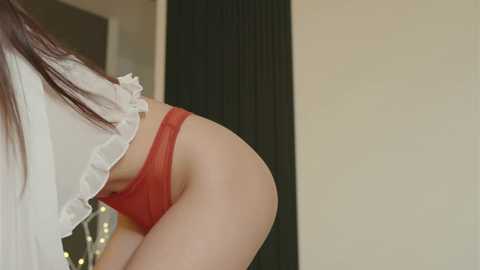 A close-up video of a woman in red lace panties, bending over, showing her round buttocks. Her long, straight, dark brown hair falls over her shoulder. The background is a plain beige wall with a dark vertical panel.