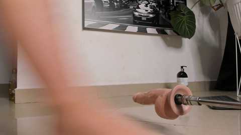 Video of a man's naked, erect penis being suctioned into a large, clear, plastic dildo. Background shows a black-and-white city street scene and a black hand lotion bottle on the floor.