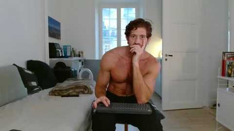 Video of a shirtless, muscular man with curly hair, sitting at a computer desk in a modern, minimalist living room.