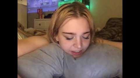 Video of a blonde woman with fair skin, wearing a gray shirt, performing oral sex on a person's penis. The background shows a TV screen displaying a blue and green image.