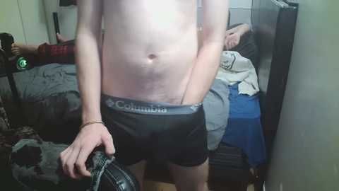 A video of a shirtless, fair-skinned person wearing Calvin Klein black boxer briefs, holding a camera, standing in a messy bedroom with a bed and clothes.