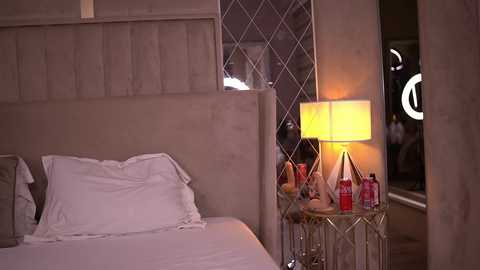Video of a modern bedroom with a beige upholstered headboard, white pillows, a white bedspread, a side table with a lamp, and a red bottle.