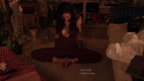 Video of a woman with long, dark hair, sitting cross-legged in a dimly lit living room, wearing a dark maroon outfit, surrounded by potted plants and a beige couch.
