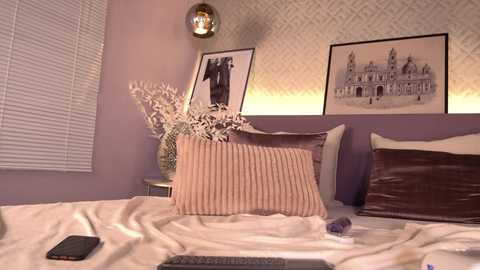 Video of a chic, lavender-painted bedroom with a white headboard, two decorative pillows, framed art, and a bedside table holding a vase with dried flowers and a smartphone.