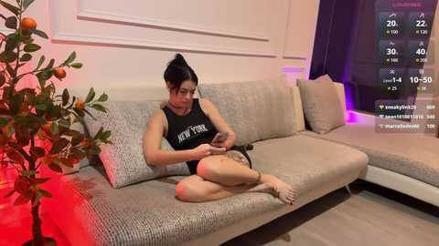 Video of a fit Asian woman with black hair in a bun, wearing a black tank top, sitting on a beige sectional sofa, playing with her phone, in a modern living room with a potted plant and a red tablecloth.