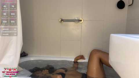 A video of a woman in a bathtub, wearing a black bra and underwear, lying on a gray towel. The bathroom has beige tiles, a metal grab bar, and a white bathtub.