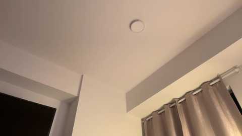 Video of a minimalist, beige-walled room with a recessed ceiling light, beige curtains, and a dark doorway. The lighting is soft and warm.