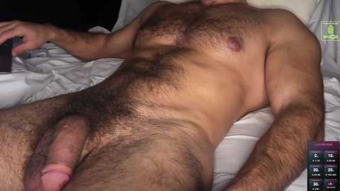 Video of a nude, light-skinned man with a hairy chest and erect penis lying on white sheets. Background includes a bed with rumpled sheets and a partially visible bedside table.