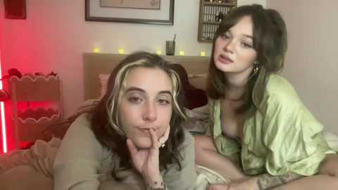 Media: Video of two young women with light skin tones and long hair, one in a green top, the other in a white top, kissing passionately on a bed, surrounded by soft, warm lighting and a framed picture.