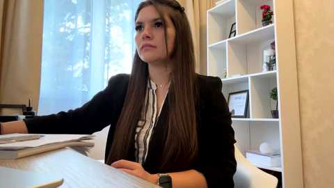 Video of a young woman with long brown hair, wearing a black blazer over a striped blouse, sitting at a desk in a well-lit office with white shelves and a window.
