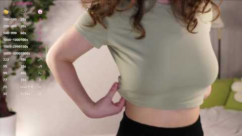Video of a woman with curly brown hair, wearing a tight, light green top and black pants, standing in a bedroom with a green sofa and a potted plant in the background.