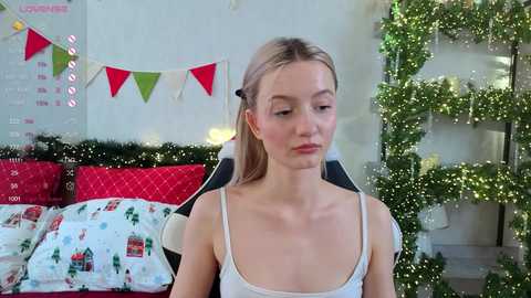 A video of a young, slender Caucasian woman with fair skin and blonde hair, wearing a white tank top, sitting in a gaming chair with festive decorations in the background.