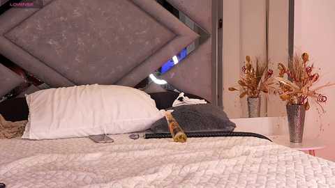 Video of a modern bedroom with a grey upholstered headboard, white bedspread, white pillows, and decorative dried flowers in a silver vase on a nightstand.