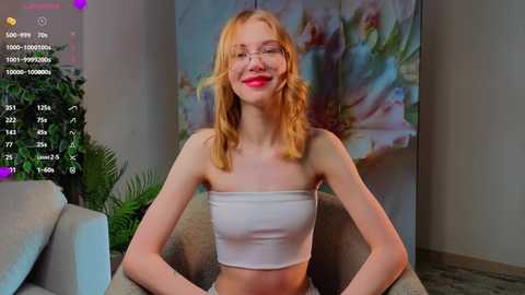 Video of a slim, fair-skinned woman with wavy blonde hair and glasses, wearing a white strapless crop top, sitting on a beige couch in a modern room with a floral painting.