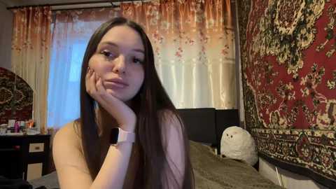 Video of a young woman with long brown hair, light skin, resting her chin on her hand, wearing a pink watch, in a cozy, intricately decorated room with red and beige floral curtains.