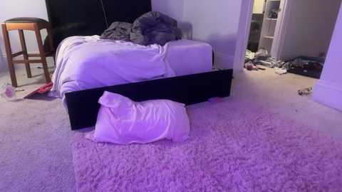 Video of a messy bedroom with a bed covered in gray and white sheets, a black headboard, and a white pillow on the floor. Clothes and shoes scattered around, carpet is light purple.