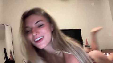 A blurry video of a young, smiling woman with long blonde hair, lying on her stomach in a dimly-lit room with a flat-screen TV in the background.