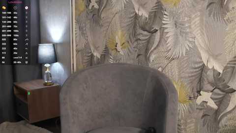 Video of a modern bedroom with a gray velvet armchair, a wooden nightstand with a black lamp, and a large wall mural depicting elegant birds and foliage in muted tones.