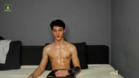 Video of a shirtless, young, muscular man with short dark hair and a prosthetic arm, sitting on a bed with white sheets, gray walls, and a black headboard.