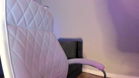 Video of a modern, white, quilted leather office chair with a swivel base against a light gray wall. A dark gray leather couch is partially visible in the background. The room has a clean, minimalist design.