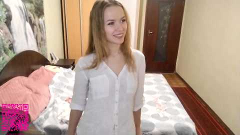 A video of a young, smiling, light-skinned woman with long blonde hair, wearing a white button-up shirt, standing in a modern bedroom with a patterned bedspread, wooden furniture, and a framed wall art.