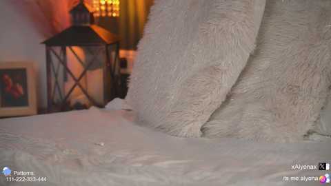 A cozy bedroom scene with a white furry blanket, a black lantern, framed artwork, and a warm, dimly lit ambiance.