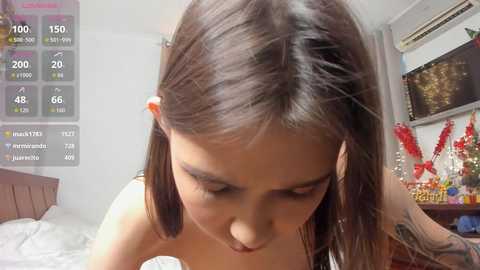 A video of a young woman with medium-length brown hair in pigtails, wearing a sleeveless top, bending over a bed in a cozy, dimly-lit room.
