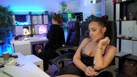 Video of a young woman with light skin, medium build, and long brown hair in a bun, wearing a black tank top, leaning on a desk in a modern, brightly lit office.