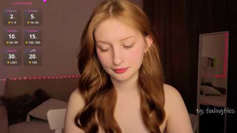 Video of a young, fair-skinned woman with long, wavy red hair, wearing a white top, sitting in a dimly lit bedroom with a bed, mirror, and purple LED strip lights in the background.