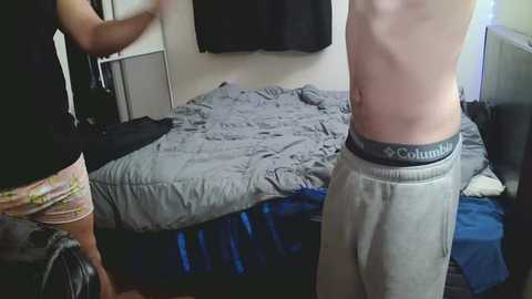 Video of a man in gray sweatpants with a black waistband, standing in a bedroom with a gray bed, a black shirt, and a black coat hanging on a wall.