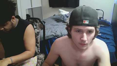Video of two shirtless young men in a messy bedroom. One man with a black cap, the other with dark hair, both seated on a bed with gray bedding and a blue blanket.