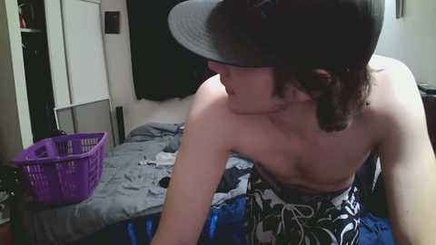 Video of a young woman with fair skin, short dark hair, and a grey cap, leaning over a bed with a blue and white patterned sheet, purple laundry basket, and a black curtain in the background.
