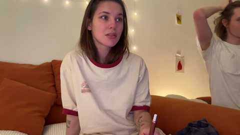 Video of a young woman with shoulder-length brown hair wearing a white T-shirt with red trim, sitting on a brown couch in a cozy room with string lights, another person partially visible.