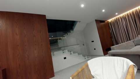 Video of a modern indoor space with dark wooden walls, white carpeted stairs, a white chaise lounge, and beige curtains; ceiling lights add warmth.