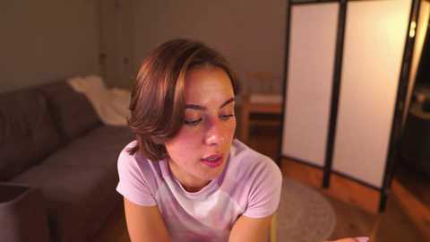 Video of a young woman with short, light brown hair, wearing a light purple t-shirt, sitting on a gray couch in a dimly lit living room with a wooden floor and folding screen.
