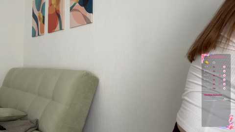 Video of a young woman with long brown hair, wearing a white top, playing a video game on a light green couch. The background features abstract art and a white wall.